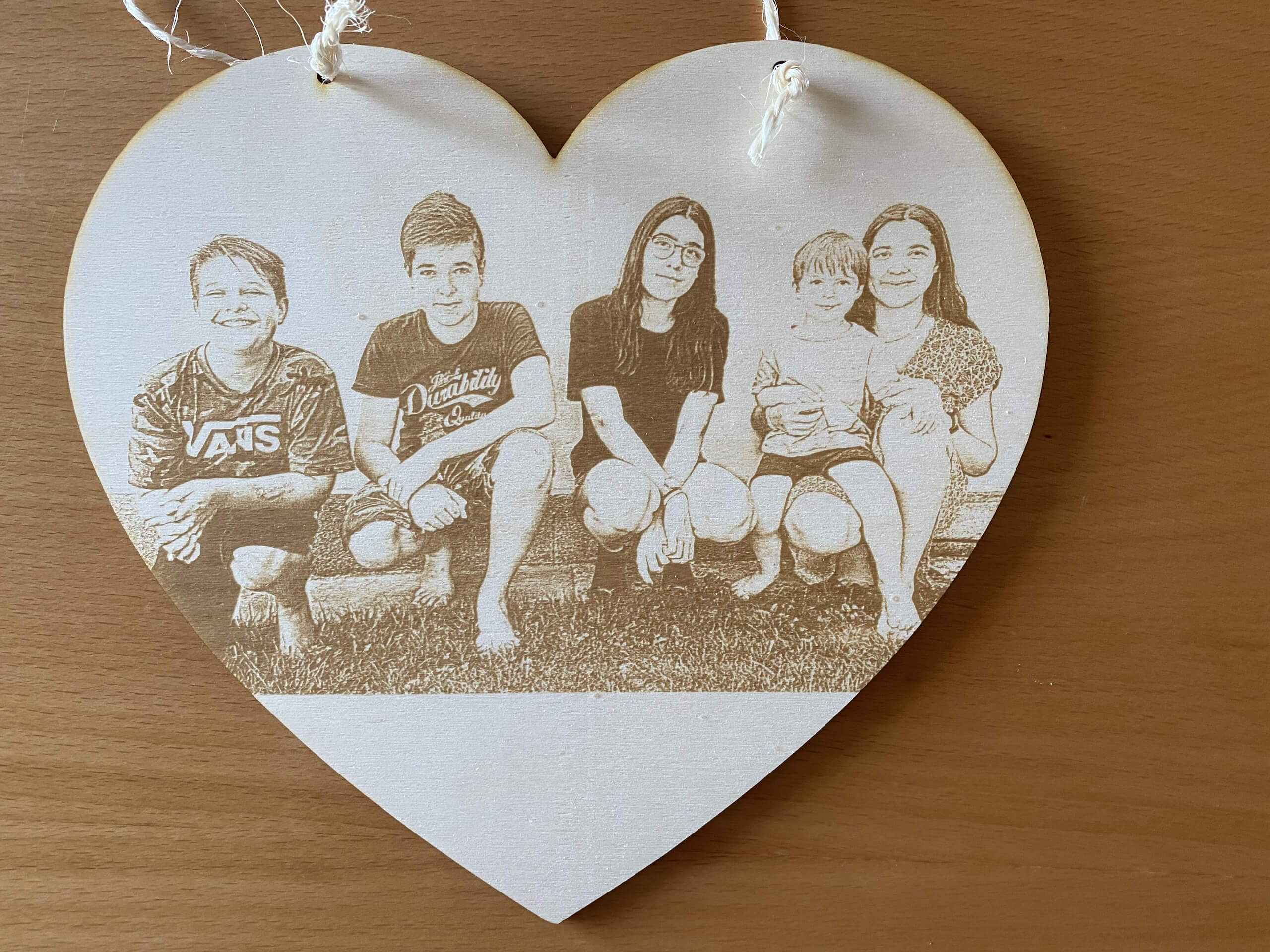 Herzförmiges Holzbild mit eingraviertem Foto von fünf lächelnden Personen auf einer Bank im Freien.