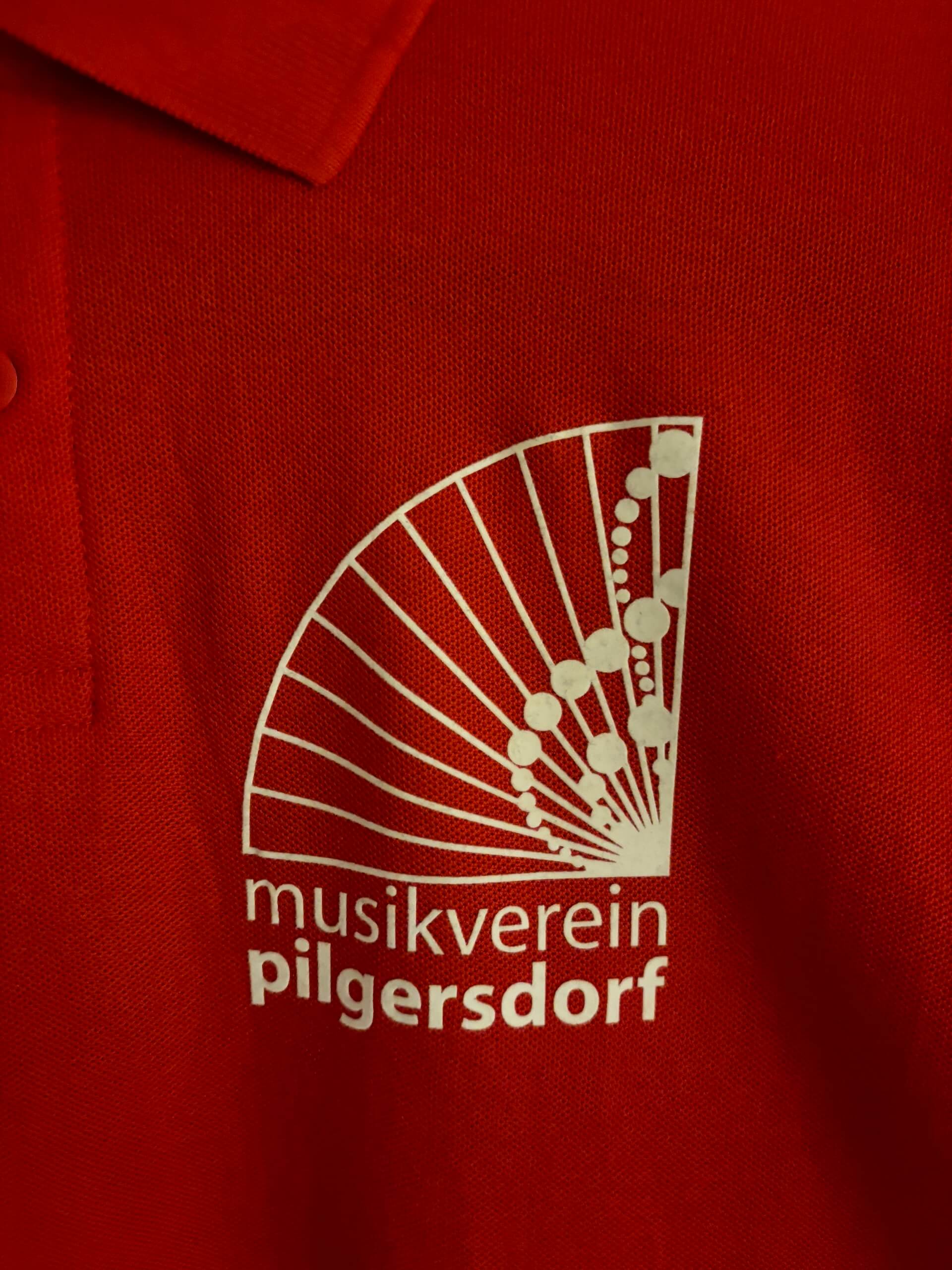 Rotes Poloshirt mit weißem Logo des Musikvereins Pilgersdorf, zeigt stilisierte Notenlinien und -noten.