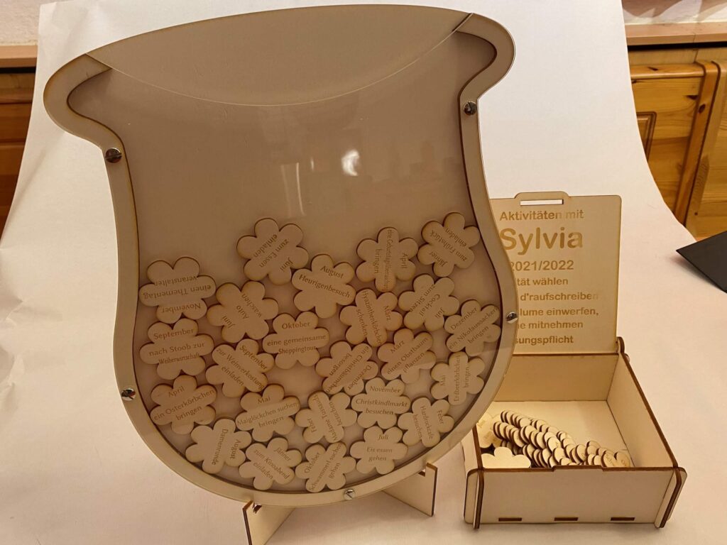 Holzdisplay mit Blumenformen für Aktivitäten mit Sylvia 2021/2022, inklusive Anweisungen zum Einwerfen und Mitnehmen.