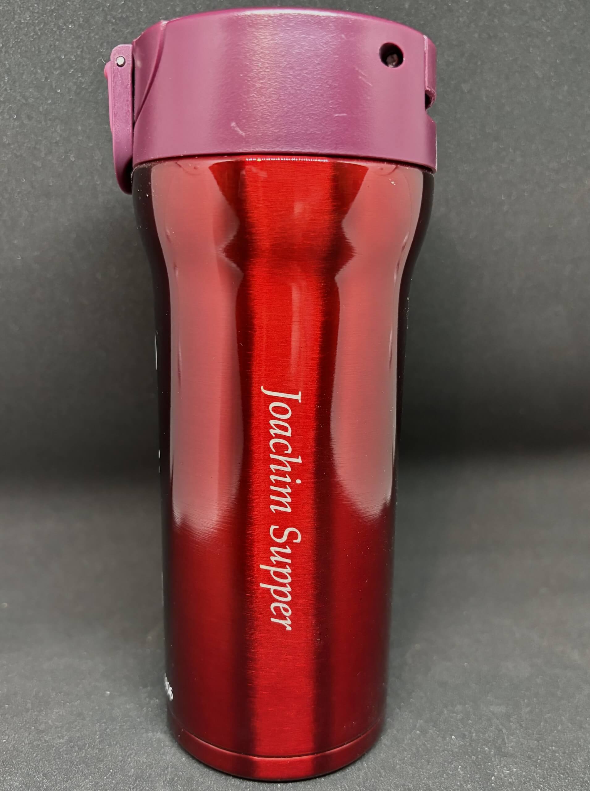 Roter Thermobecher mit lila Deckel und graviertem Namen, vor schwarzem Hintergrund stehend.