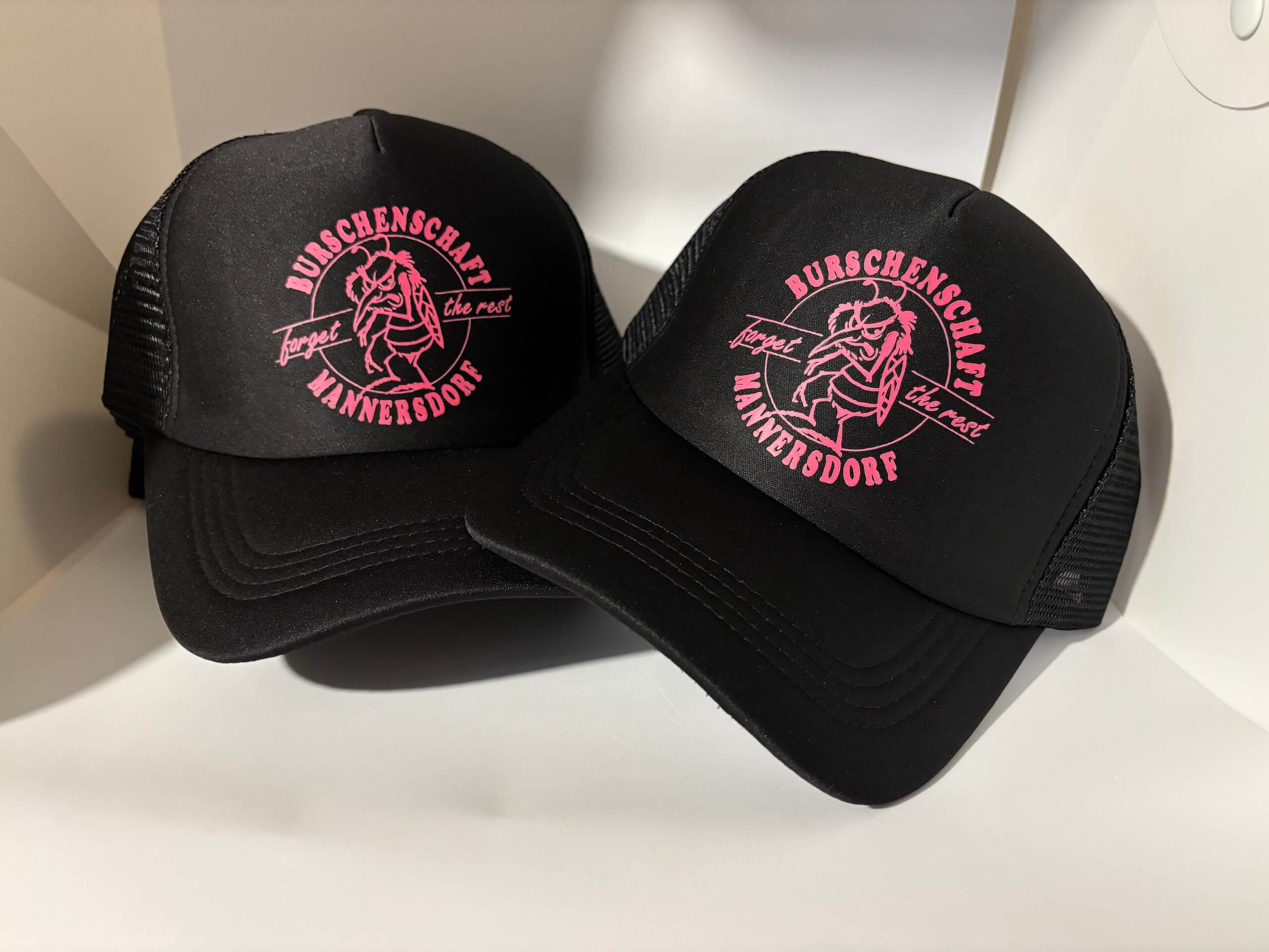Schwarze Basecaps mit pinkem Burschenschaft Mannersdorf Logo auf weißem Hintergrund. Trucker-Design.