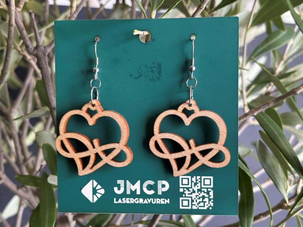 Holzohrringe mit Herzdesign, präsentiert auf einer Karte von JMCP Lasergravuren, im Hintergrund grüne Blätter.