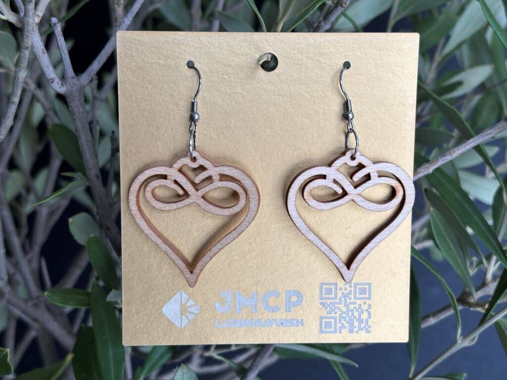 Holz-Ohrringe in Herzform mit Unendlichkeitssymbol, präsentiert auf JMCP Lasergravuren-Verpackung vor Pflanzenhintergrund.