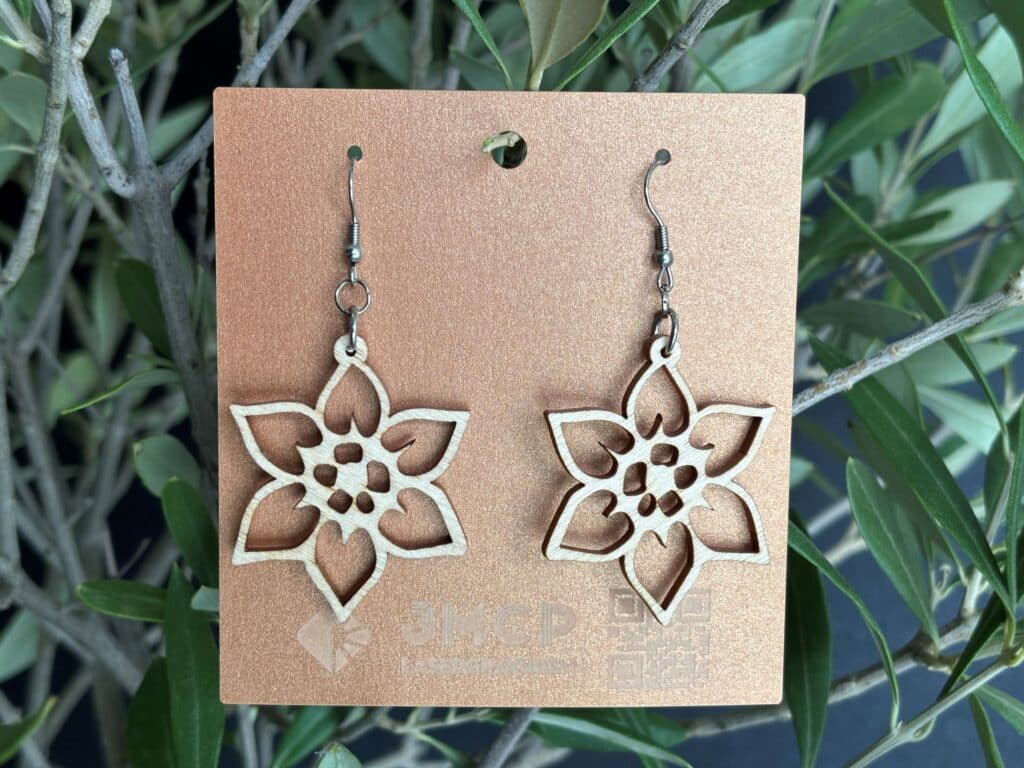 Lasergravierte Ohrringe in Blütenform auf einer Karte, präsentiert vor grünem Pflanzenhintergrund.