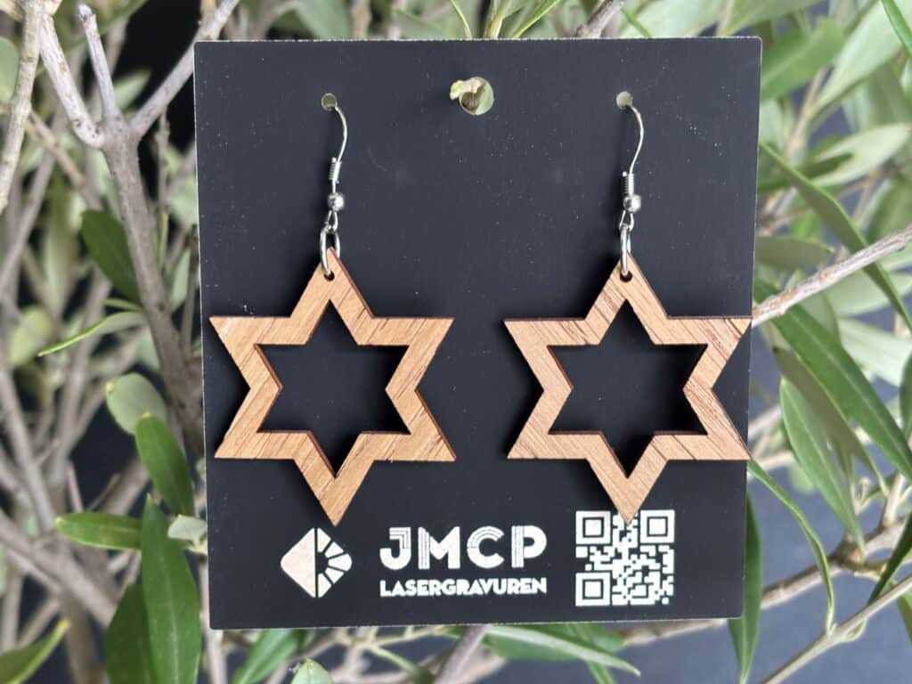 Holzohrringe in Sternform auf schwarzem JMCP-Kärtchen, platziert vor grünen Blättern, präsentieren Lasergravuren.