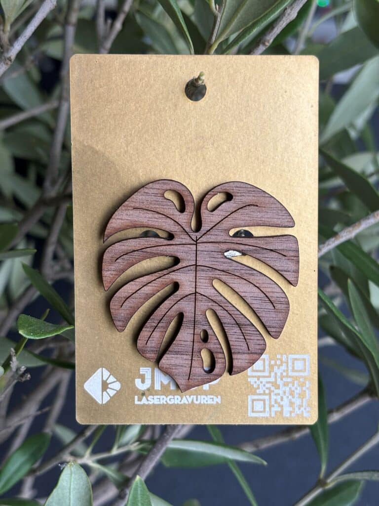 Holzgeschnitztes Monstera-Blatt, gestaltet von JMCP Lasergravuren, auf goldener Karte vor Pflanzenhintergrund.