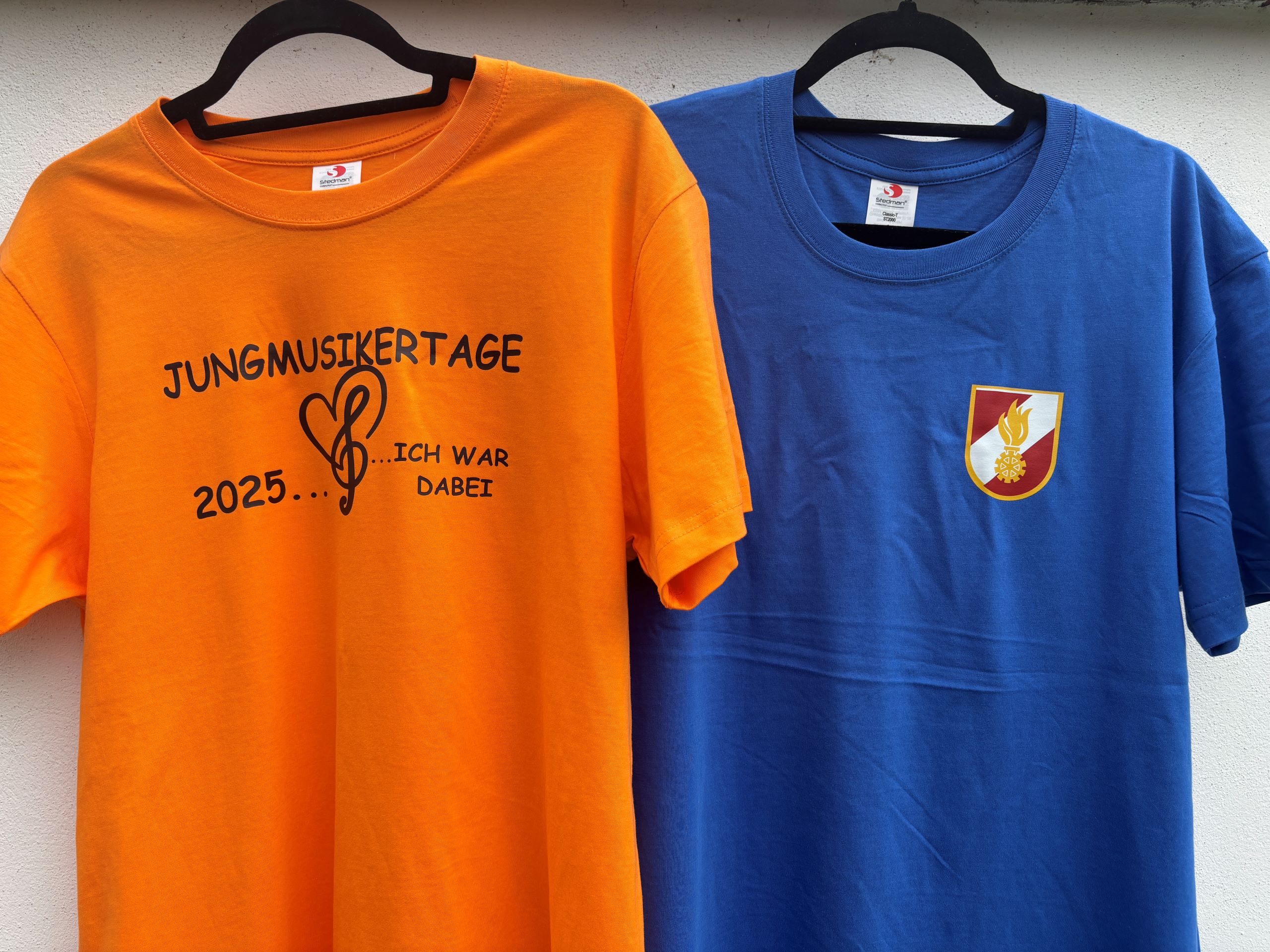 Orange und blaues T-Shirt mit Jungmusikertage 2025 und Logo, auf Kleiderbügeln vor weißem Hintergrund.