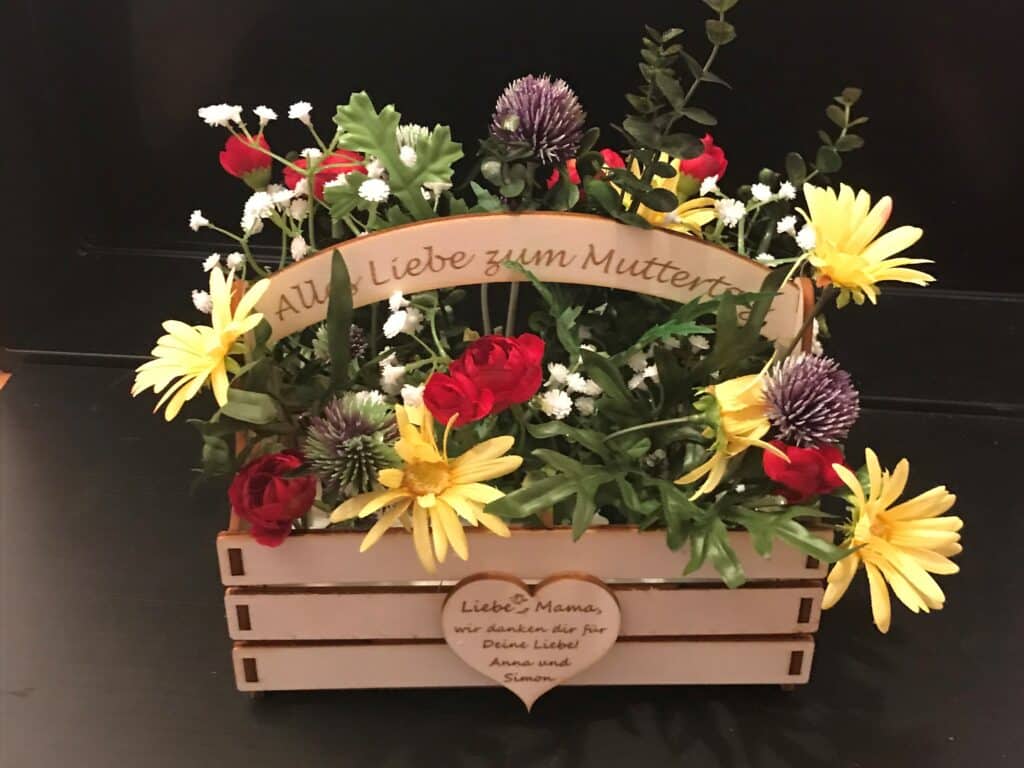 Blumengesteck in Holzkiste mit lasergraviertem Muttertagsgruß und personalisierter Botschaft in Herzform auf der Vorderseite.