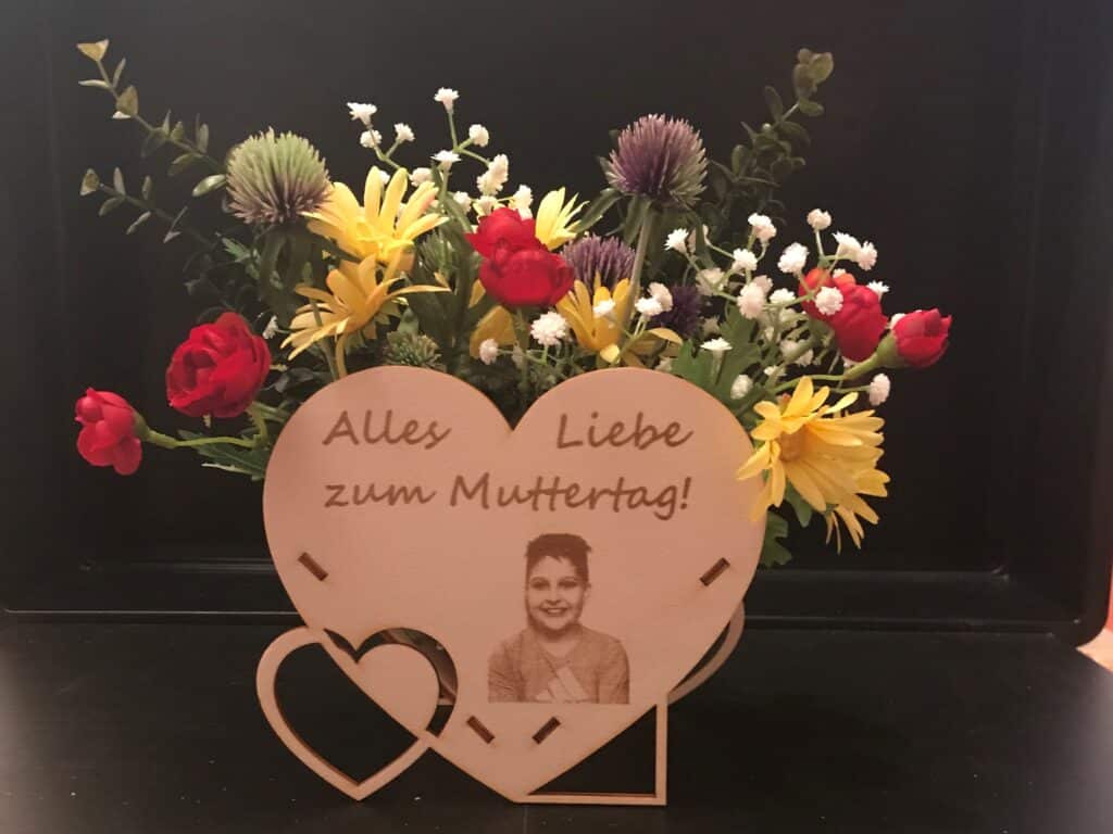 Herzförmige Holzkarte mit „Alles Liebe zum Muttertag!“ vor einem bunten Blumenstrauß als personalisiertes Geschenk.