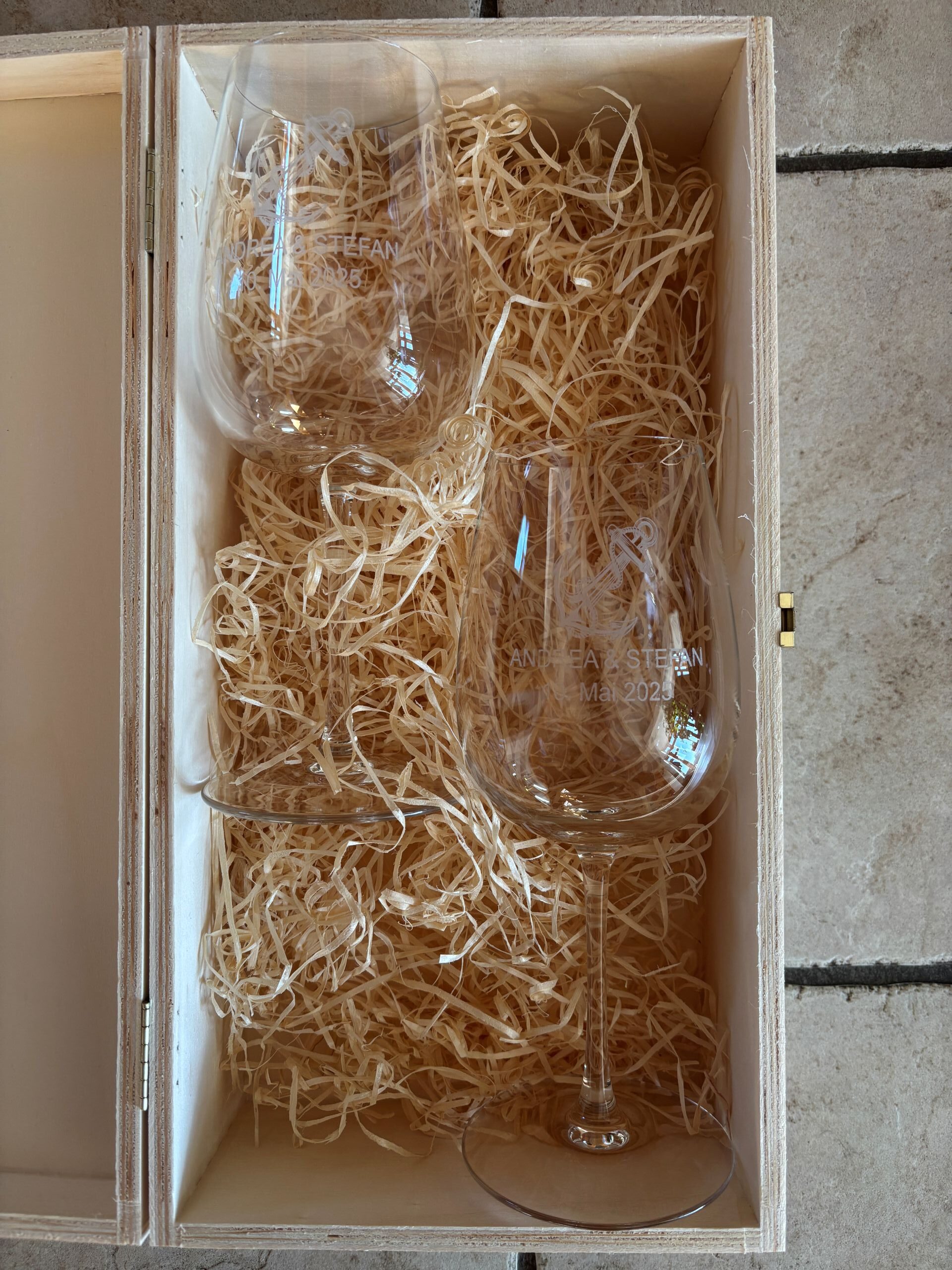 Personalisierte Weingläser in Holzkiste mit Gravur, umgeben von Holzwolle, ideal als Hochzeitsgeschenk.