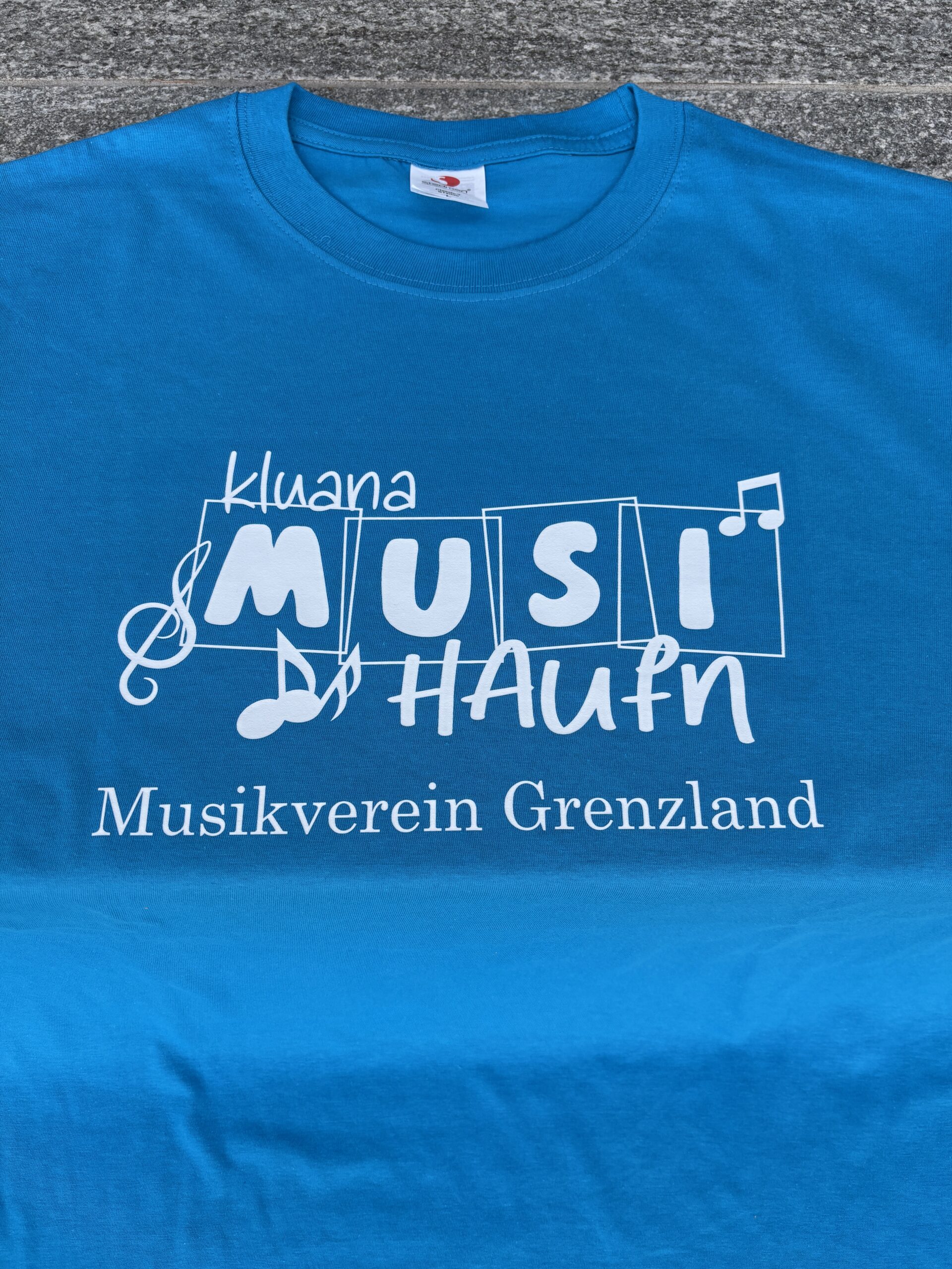 Blaues T-Shirt mit „Musi Hau'n“ und „Musikverein Grenzland“ Schriftzug, ideal für kulturelle Veranstaltungen.