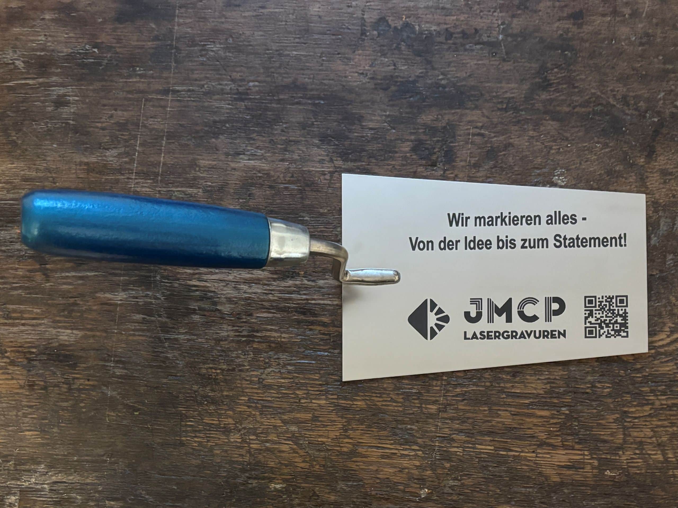 Werkzeug mit blauem Griff neben JMCP Lasergravuren-Karte auf Holzoberfläche, bietet Gravurdienstleistungen an.