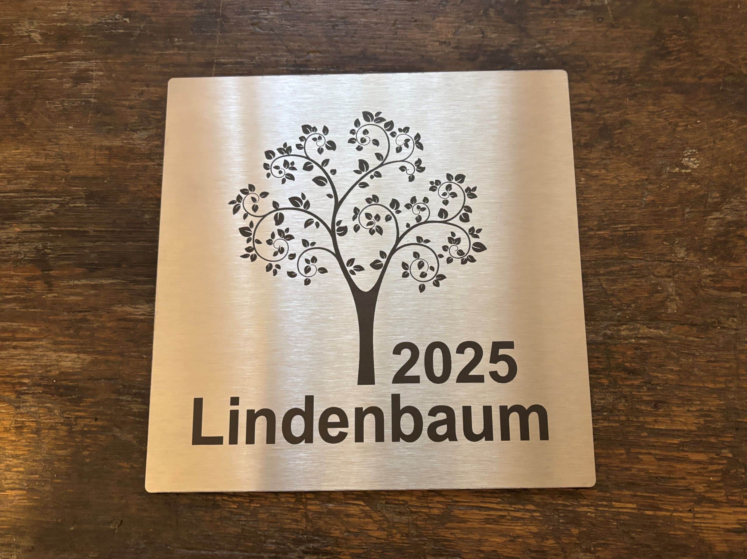 Edelstahlplatte mit lasergraviertem Baum und Inschrift 2025 Lindenbaum auf holzartigem Hintergrund.