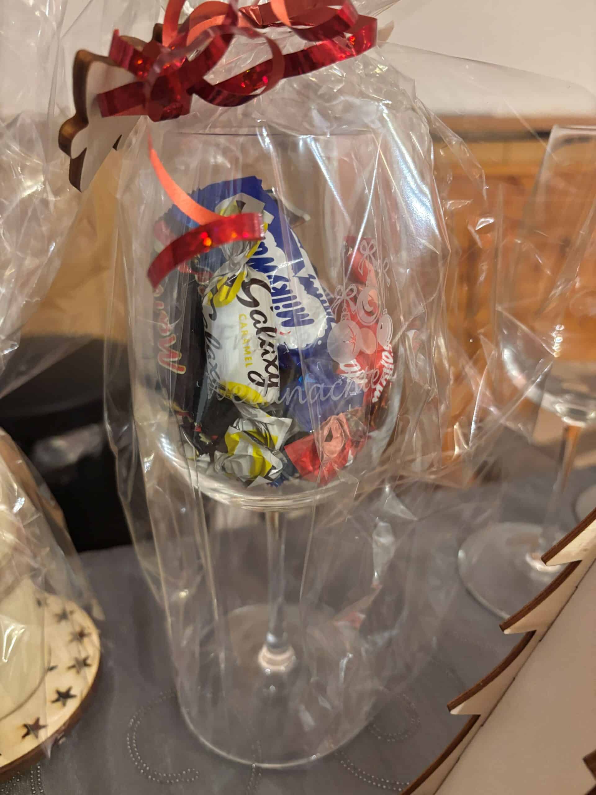 Weinglas mit Süßigkeiten gefüllt und in Geschenkfolie verpackt, dekoriert mit rotem Band und Holzstern.