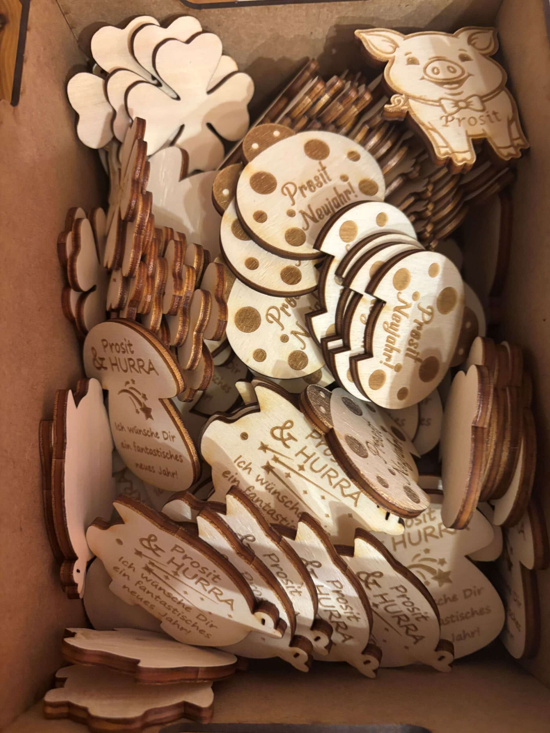 Holzgeschenke mit Gravuren „Prosit Neujahr!“ und „Prosit & Hurra“, darunter Glückssymbole wie Schweinchen und Kleeblätter.