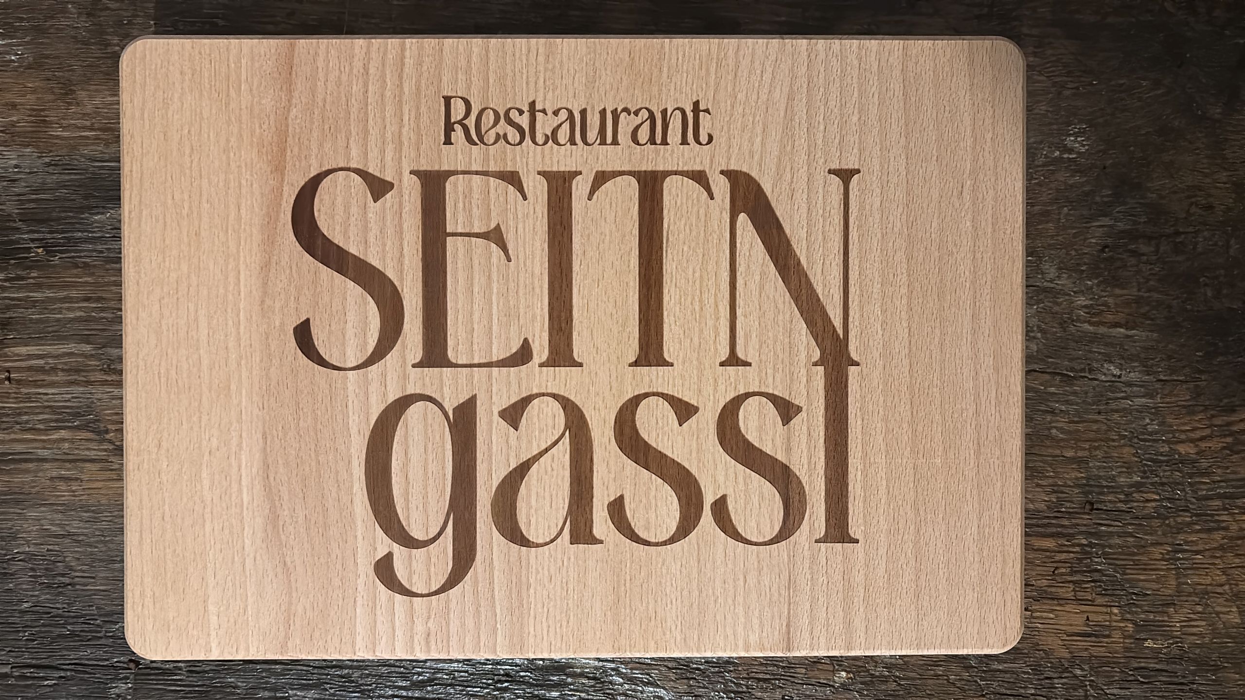 Holzschild mit Lasergravur, Restaurant SEITN gassi, auf rustikalem Holztisch liegend.