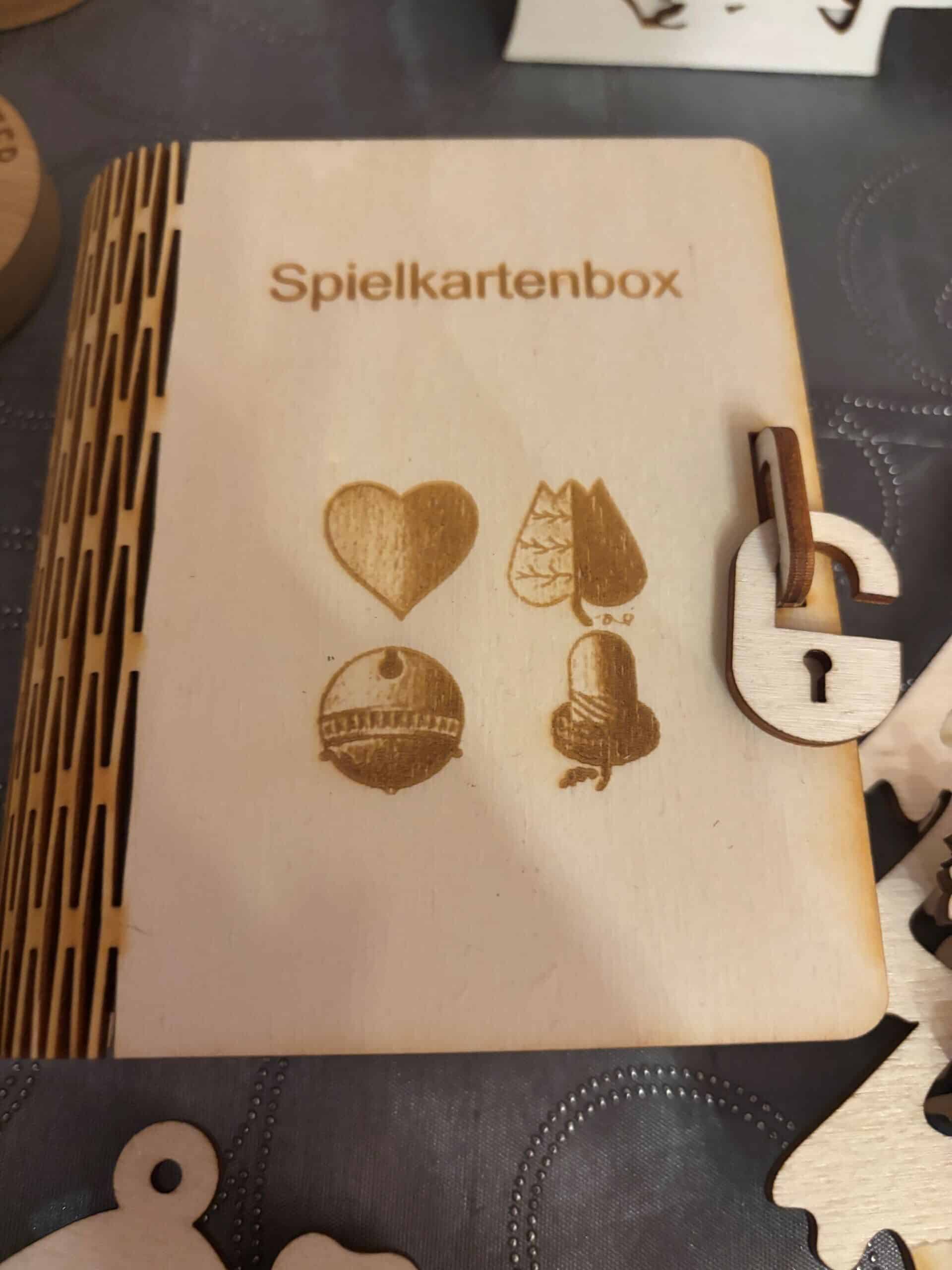 Holzspielkartenbox mit lasergravierten Kartensymbolen und einem Schlossverschluss auf dunkler Tischdecke von JMCP Lasergravuren.