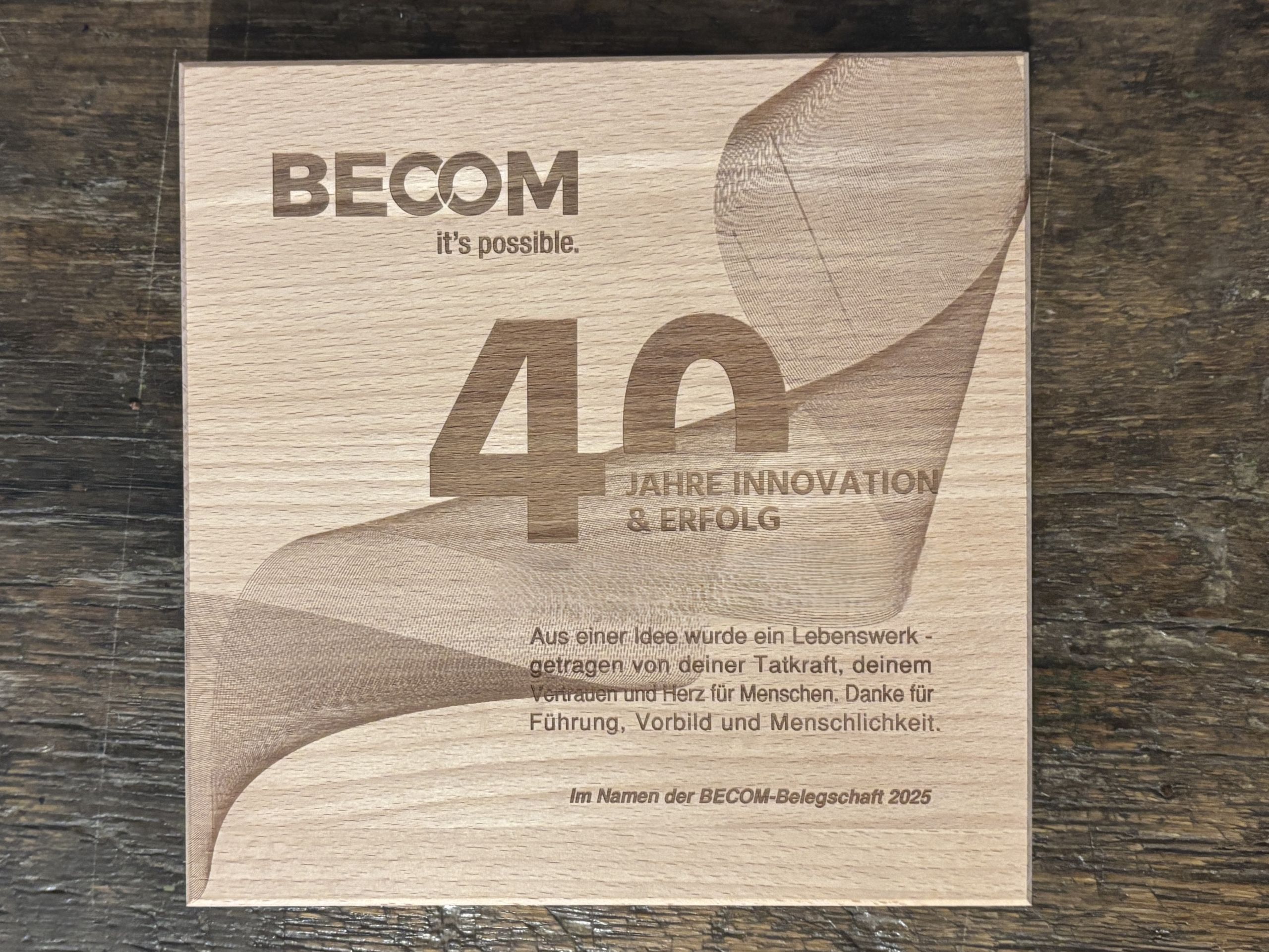 Holztafel mit Lasergravur „BECOM - 40 Jahre Innovation & Erfolg“, auf einer dunklen Holzoberfläche.
